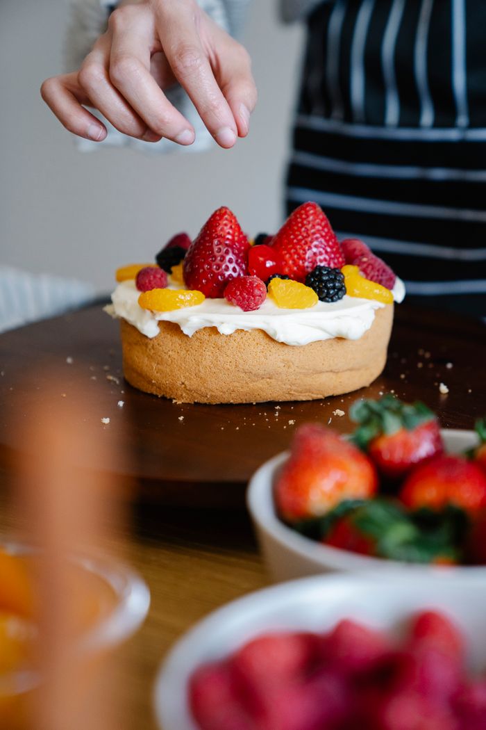 Close-up of a fresh fruit-topped cake being finished with a hand adding berries.